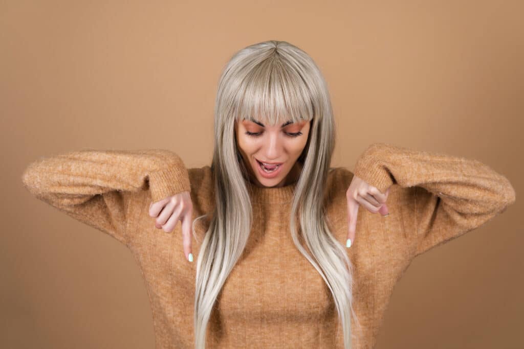 Blonde girl with bangs and brown daytime makeup in a sweater on a beige background smiles excitedly points her finger down